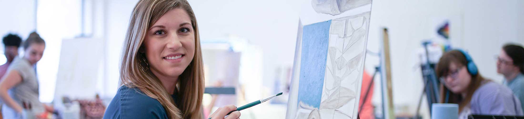 Student holding paint brush with painting in front of her in art class.