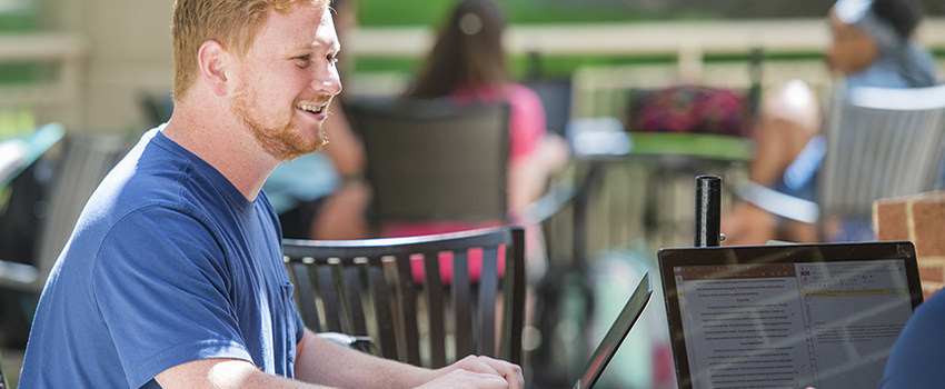 Male student working on laptop at table outside