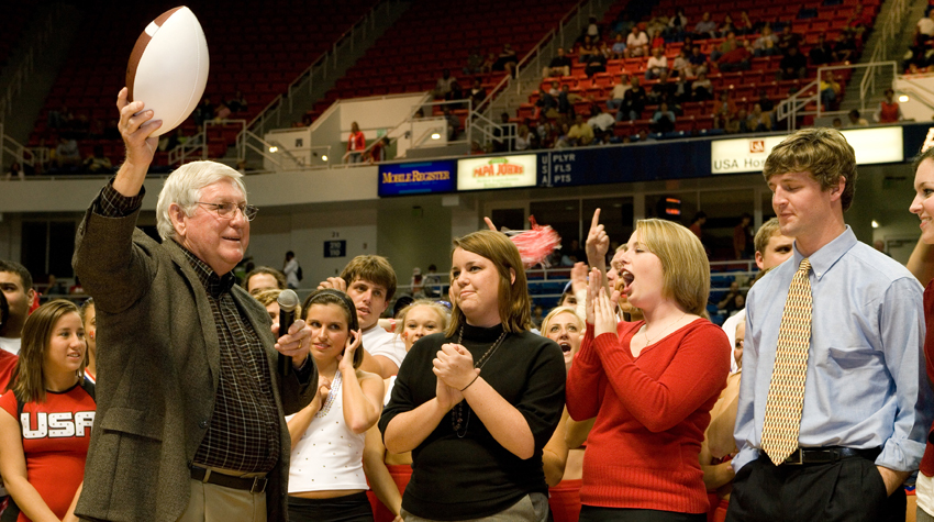 President Moulton holding football in the air in the Mitchell Center with crowd cheering.