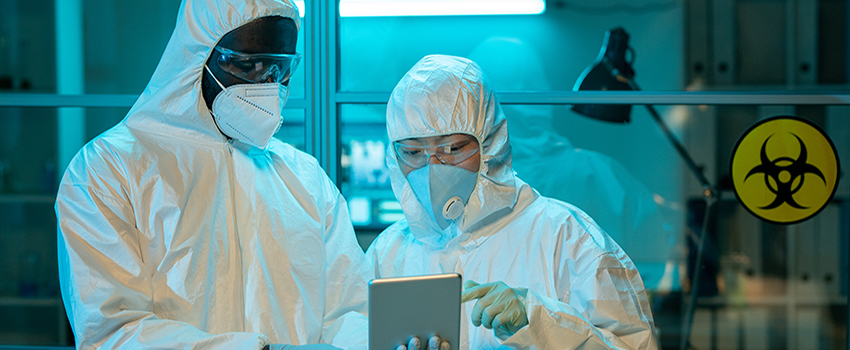 Two people in hazmat suits in lab looking at tablet.